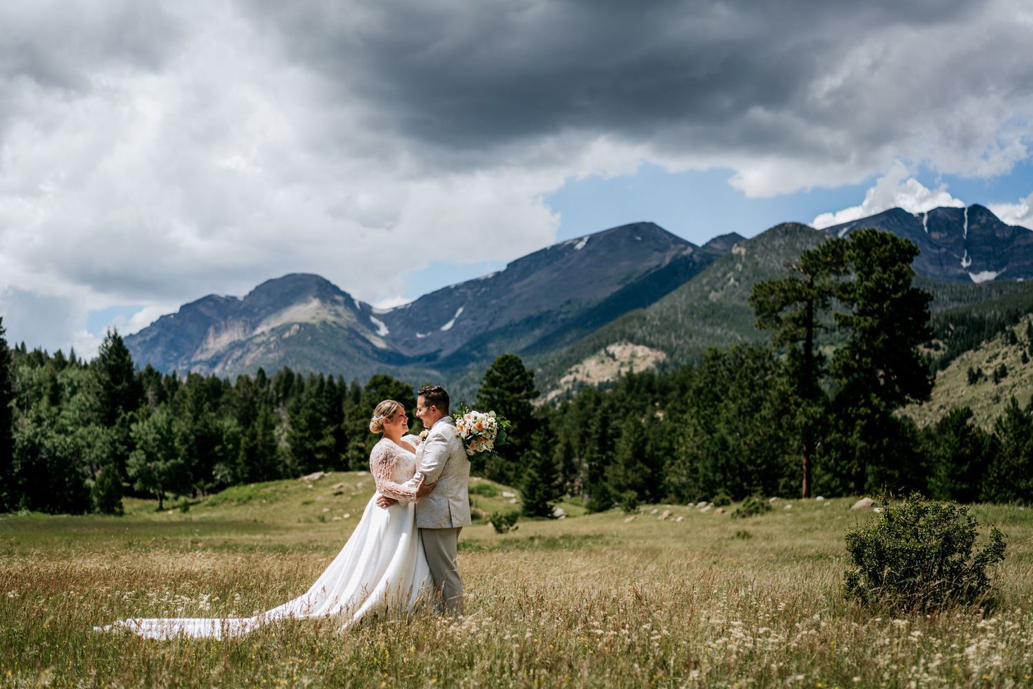 Couple embracing in a mountain field with dramatic storm clouds and peaks in the background.