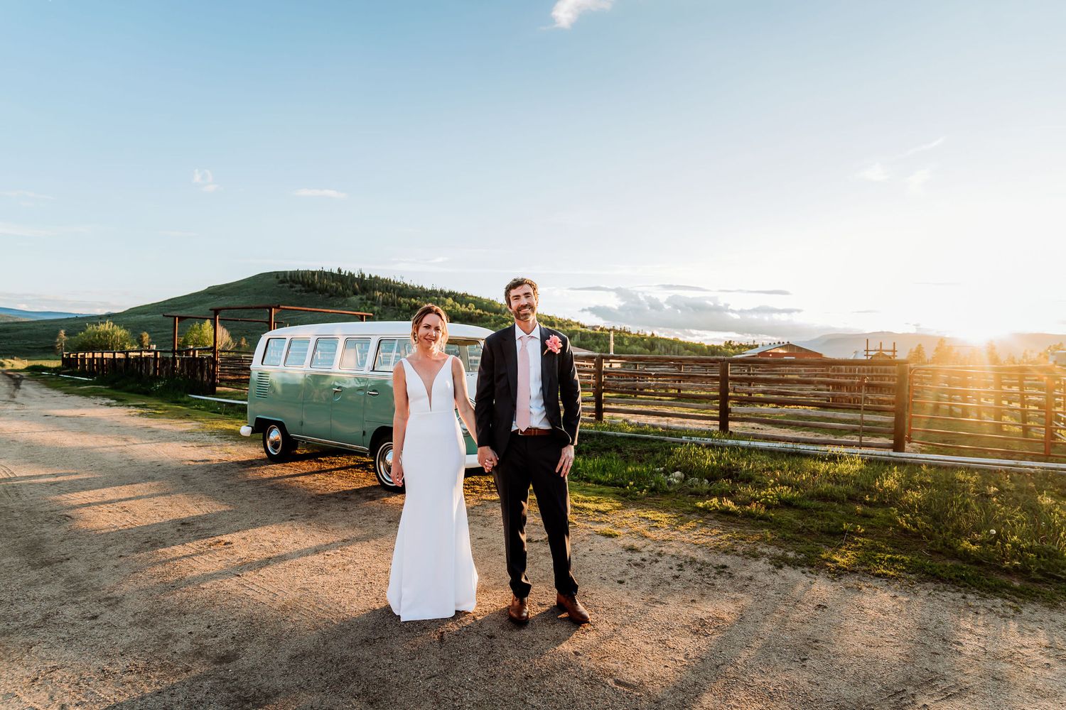 A couple poses in front of a classic VW bus on a scenic mountain road at golden hour.