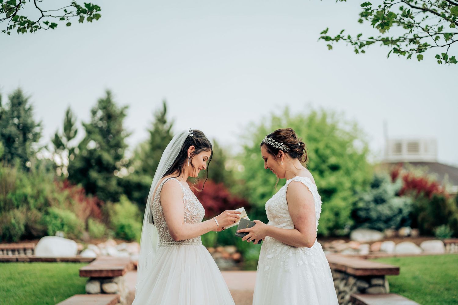 Two people in white wedding dresses share a tender moment in a garden setting.