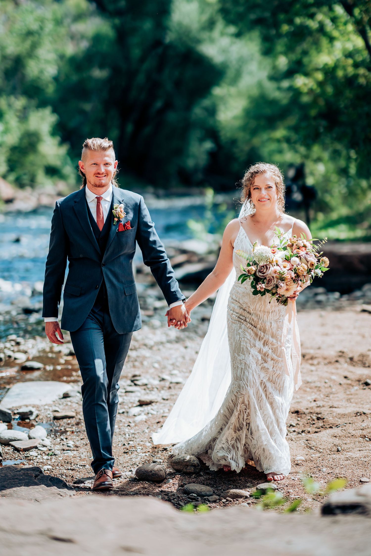 Newlyweds walk hand in hand along rocky riverbank path with flowing water and cliffs.