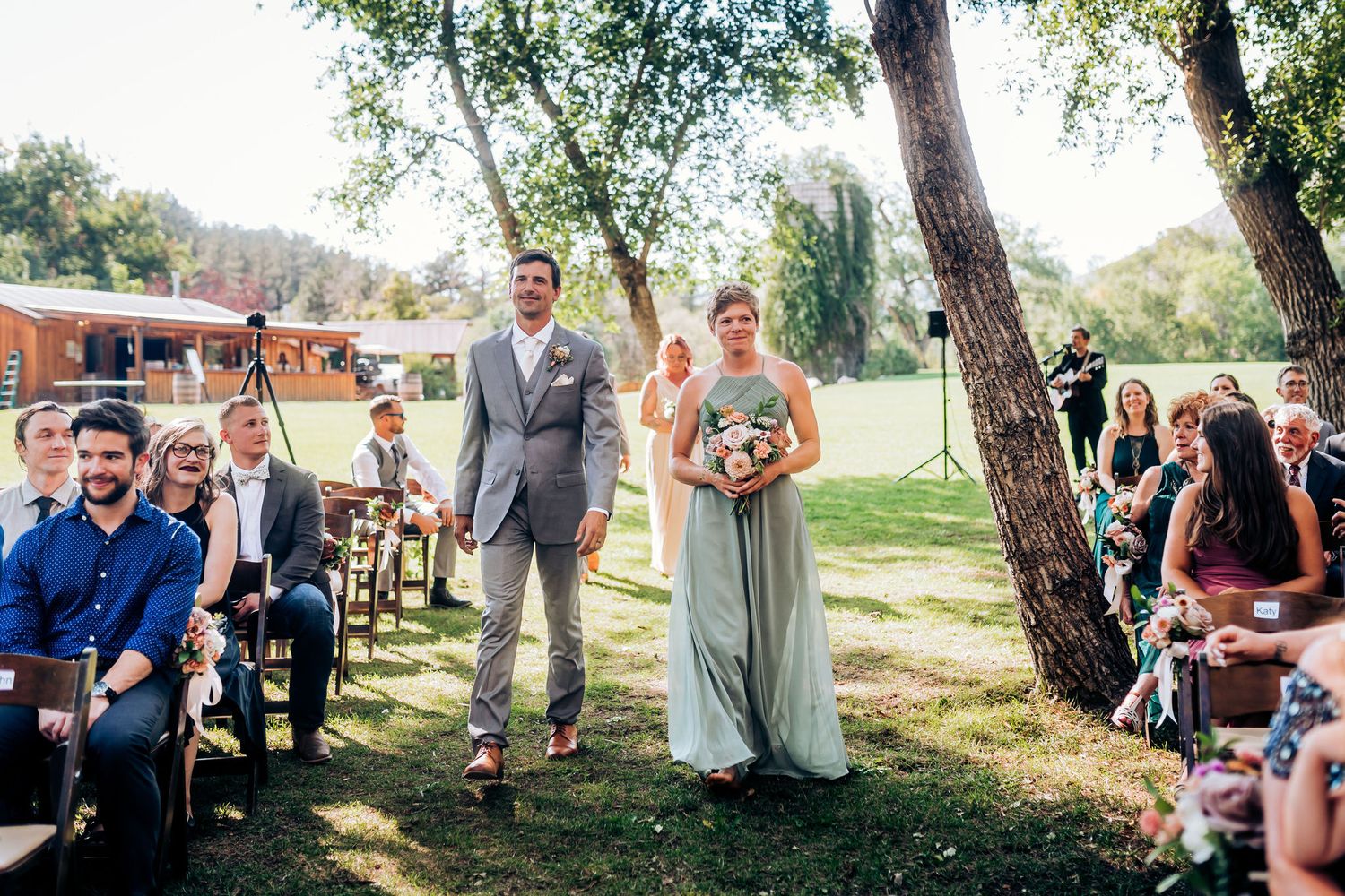 Wedding ceremony in progress with guests seated on wooden chairs in an outdoor garden setting.