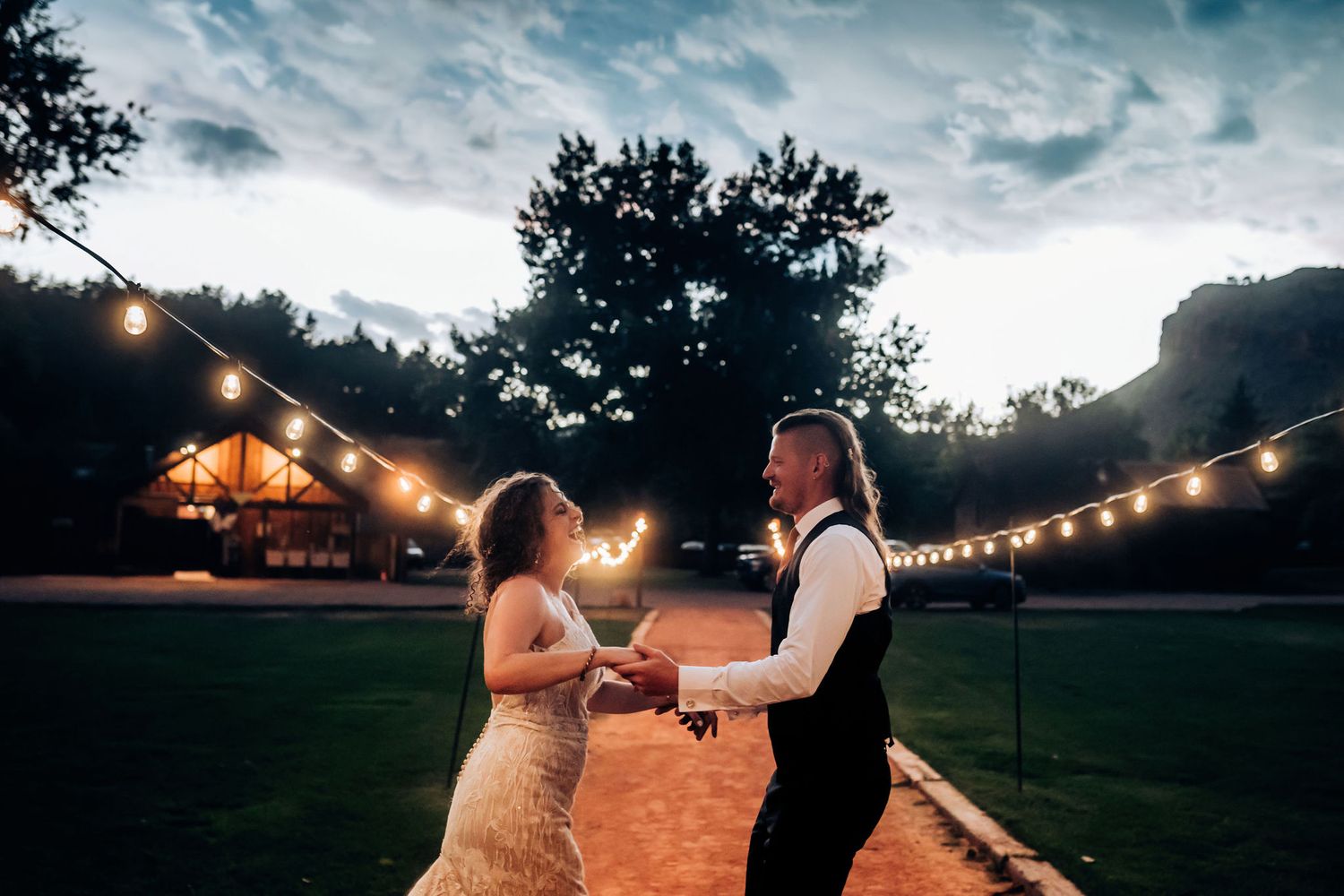 Couple dances under string lights at dusk with a rustic barn venue in the background.
