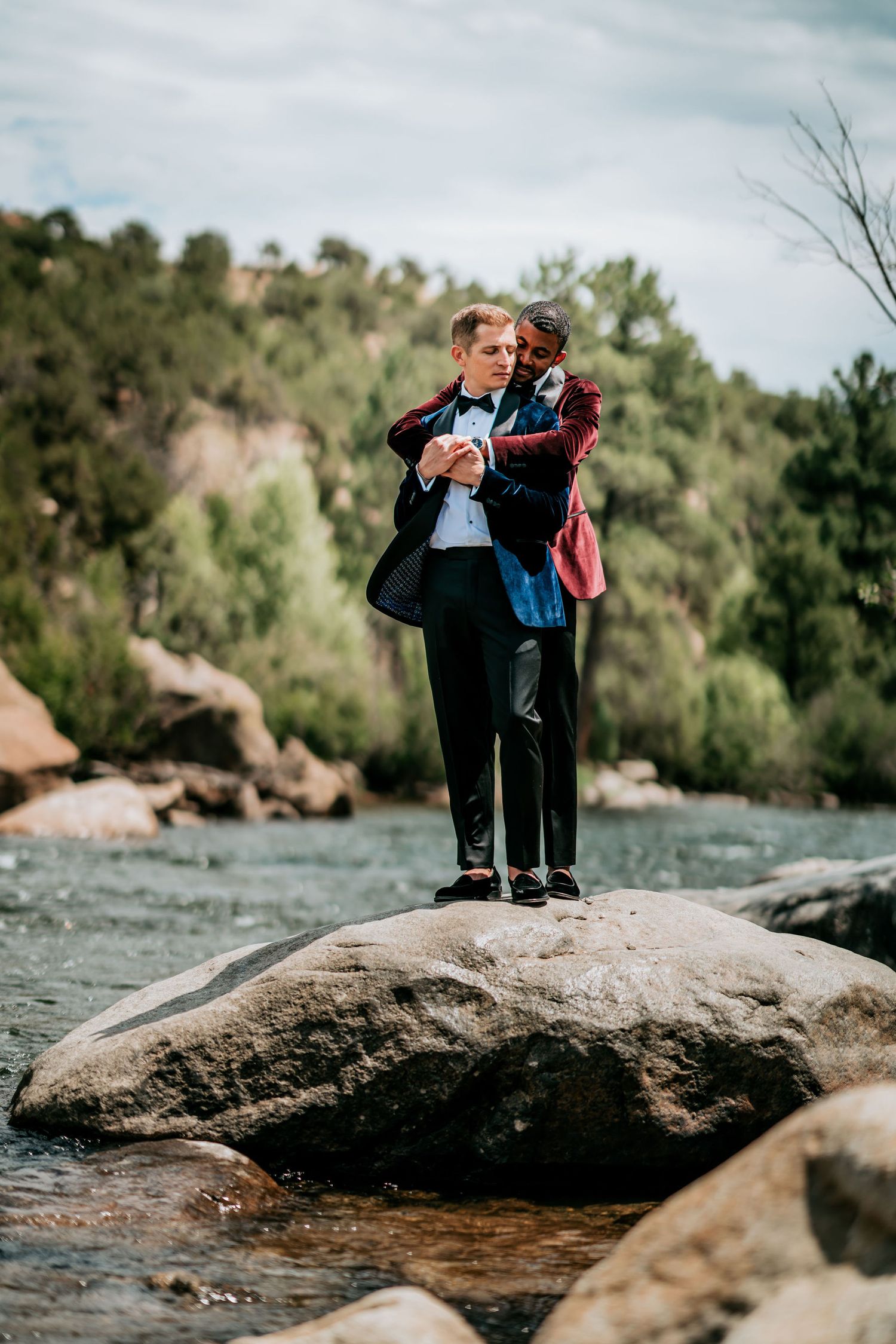 A romantic moment between two people in formal wear standing on a large rock by a river.