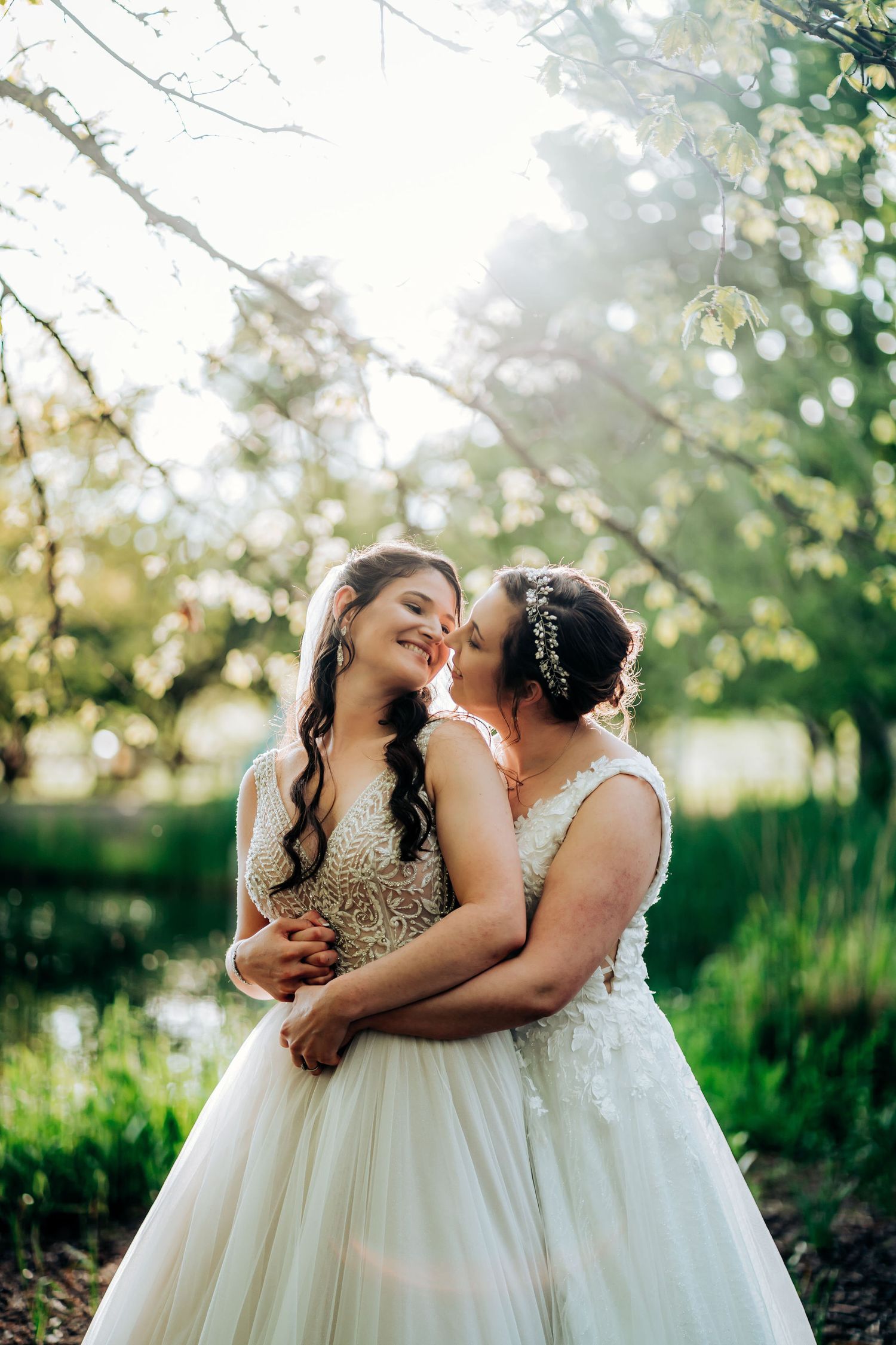 Two brides in white dresses share an intimate moment in a sun-dappled garden setting.