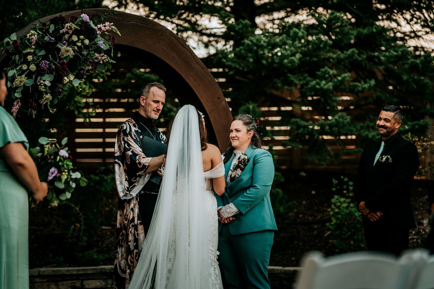 Two brides smile at each other during outdoor wedding ceremony beneath floral circular arch with officiant present.