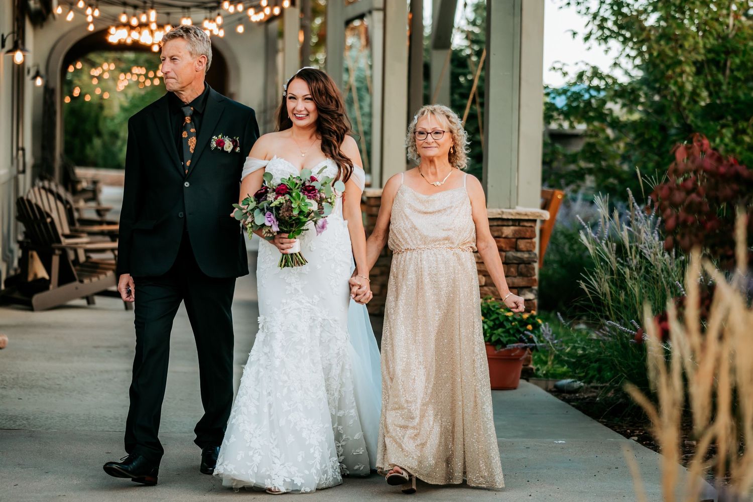 Bride in white lace gown walks down aisle with parents at outdoor wedding ceremony.