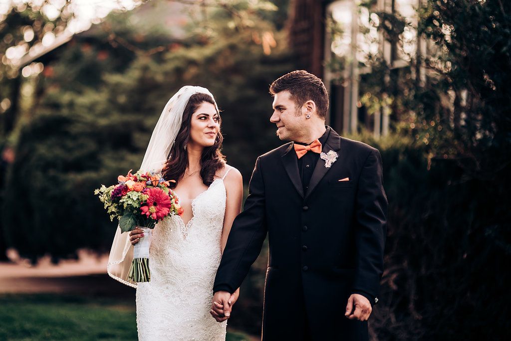 A romantic garden wedding portrait with a bride in white lace holding vibrant red flowers next to her groom in black.