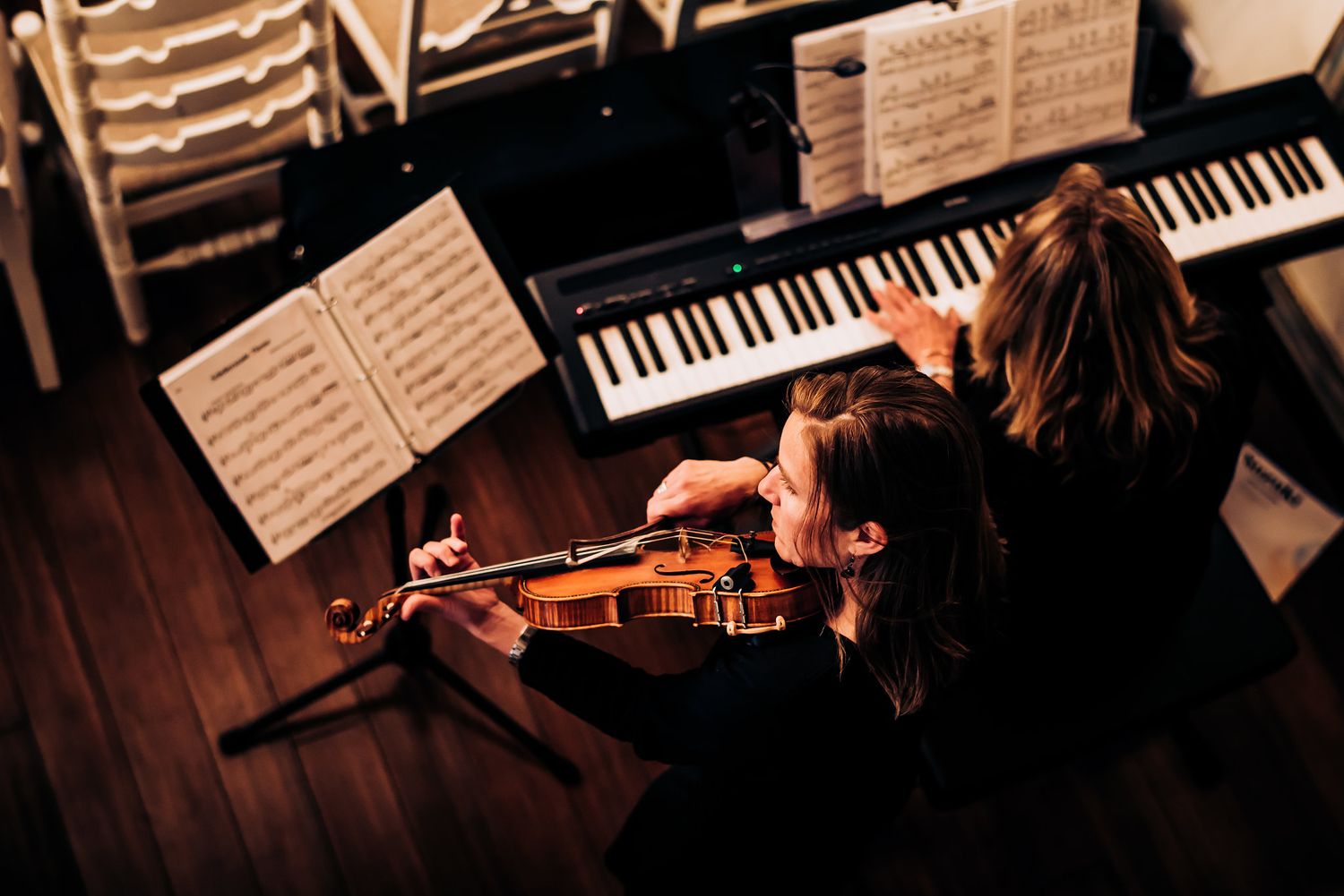 Musician playing violin next to piano with sheet music in dimly lit room.