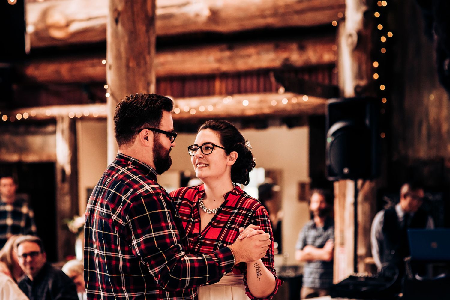 A couple wearing matching plaid shirts share a romantic dance in a rustic barn setting.
