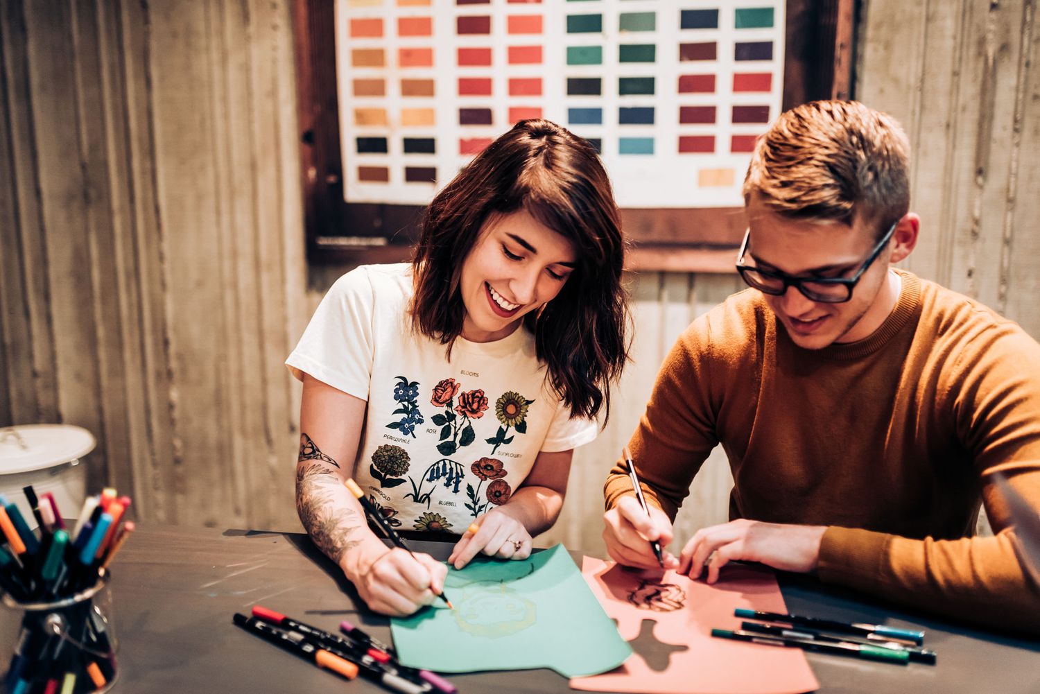 Two artists work together on a creative project at a desk with art supplies.