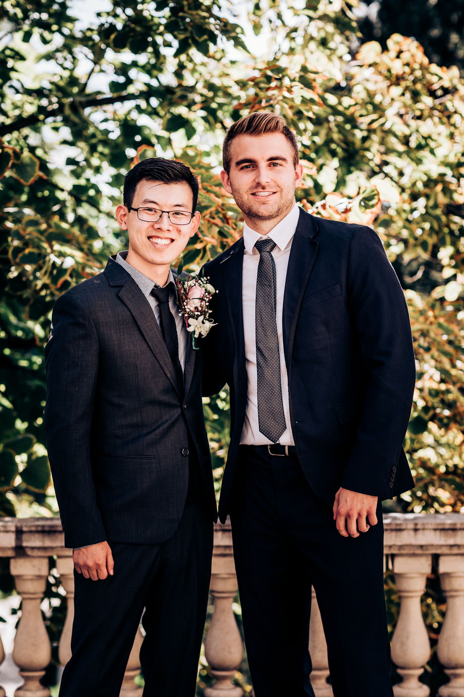 Two groomsmen in dark suits posing together at an outdoor venue.