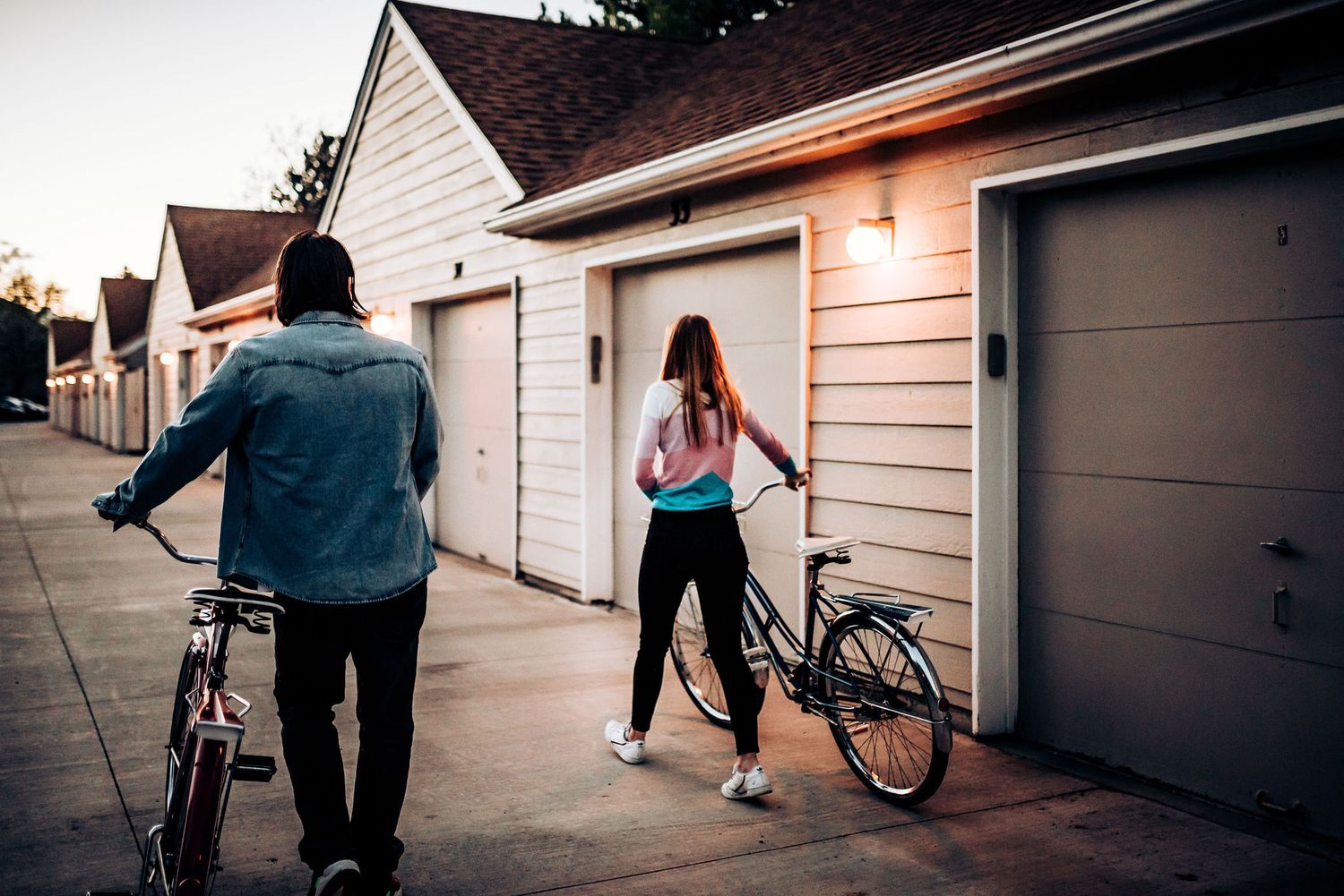 A casual couple with bicycles stands near garage doors at dusk.