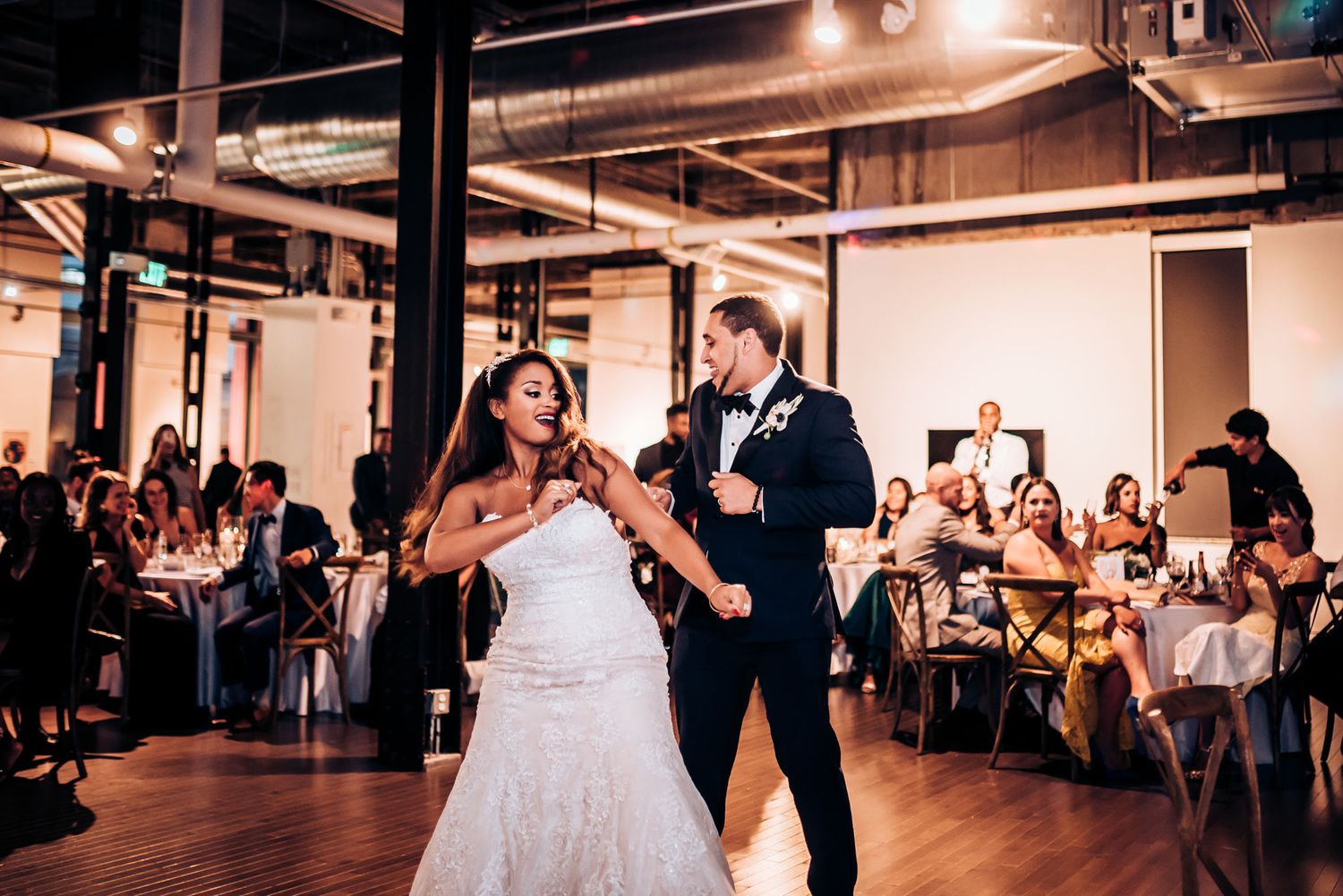 A couple shares their first dance on a wooden floor while wedding guests capture the moment on their phones.