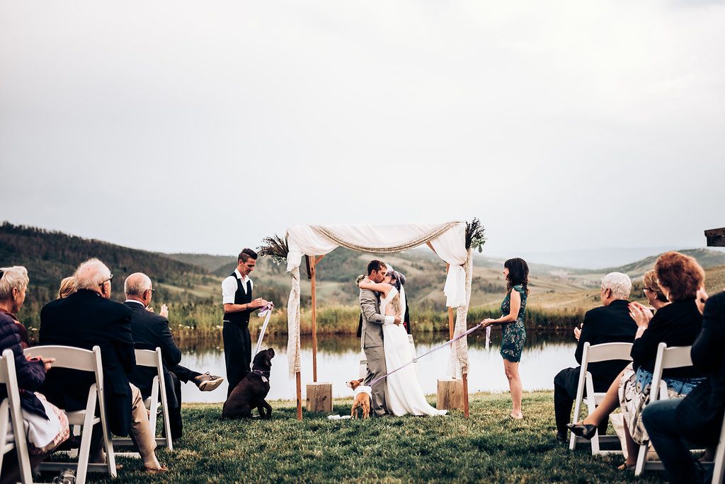 Outdoor wedding ceremony setup with white chairs facing a fabric-draped arch beside a peaceful lake.