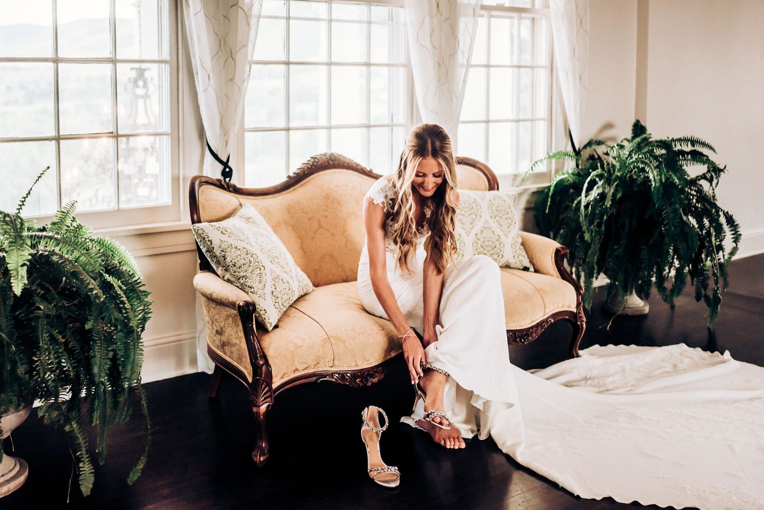 Elegant vintage sofa scene with a white wedding dress and indoor plants by large windows.