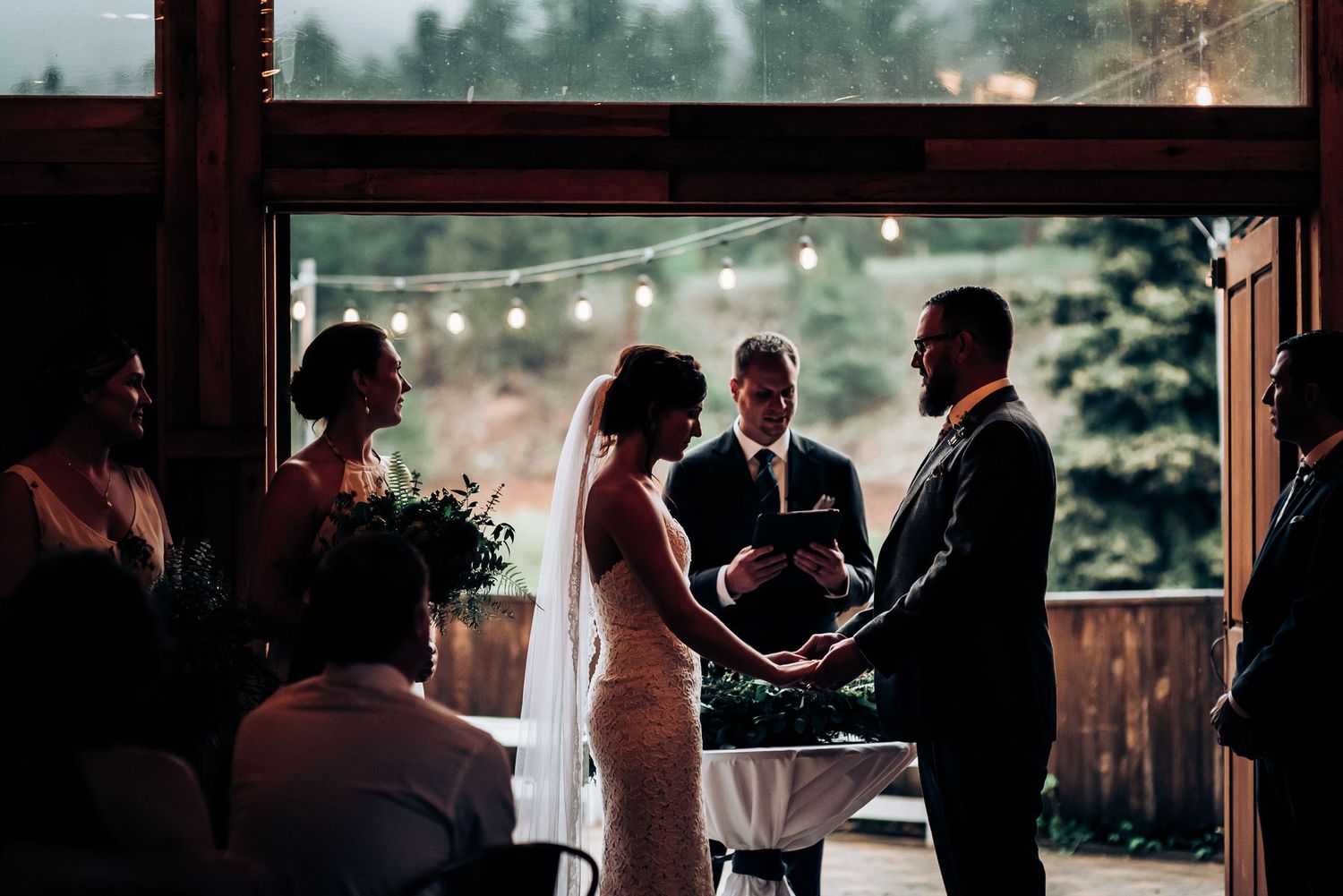 A wedding ceremony in progress inside a rustic venue with string lights visible through windows.
