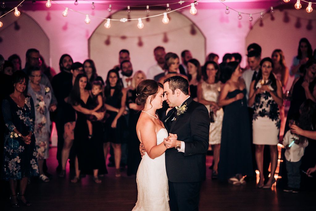 A couple shares their first dance at a wedding reception under pink lighting while guests watch from the dance floor.