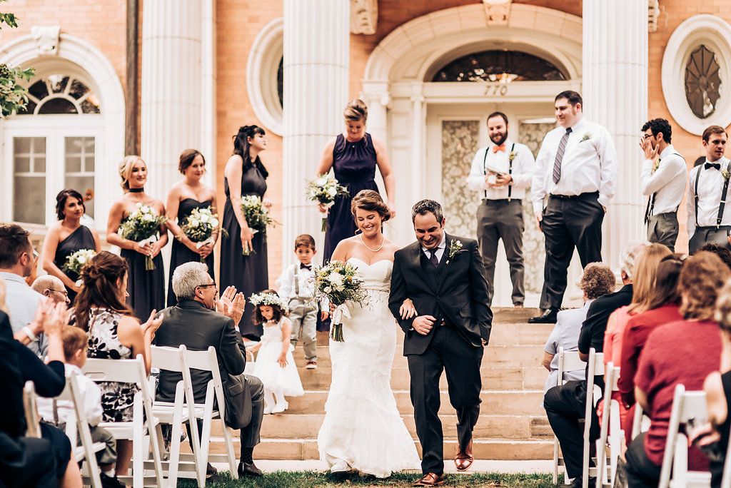 Newlyweds walk down steps after ceremony while guests seated on white chairs look on.