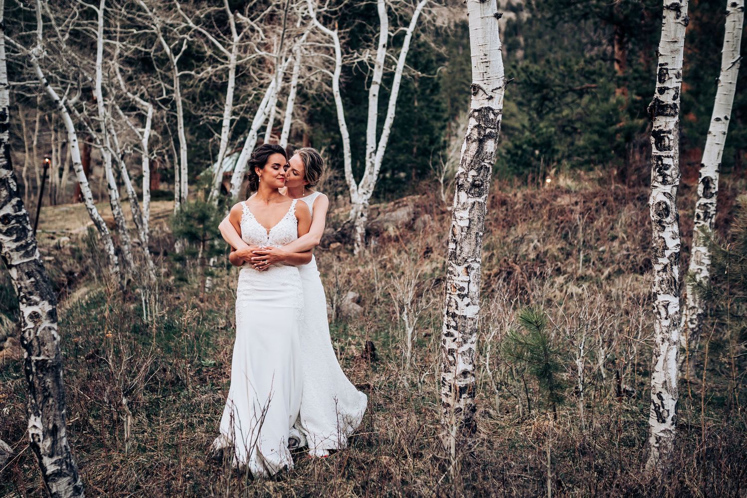 Two people in white wedding gowns embrace in a serene birch forest setting.