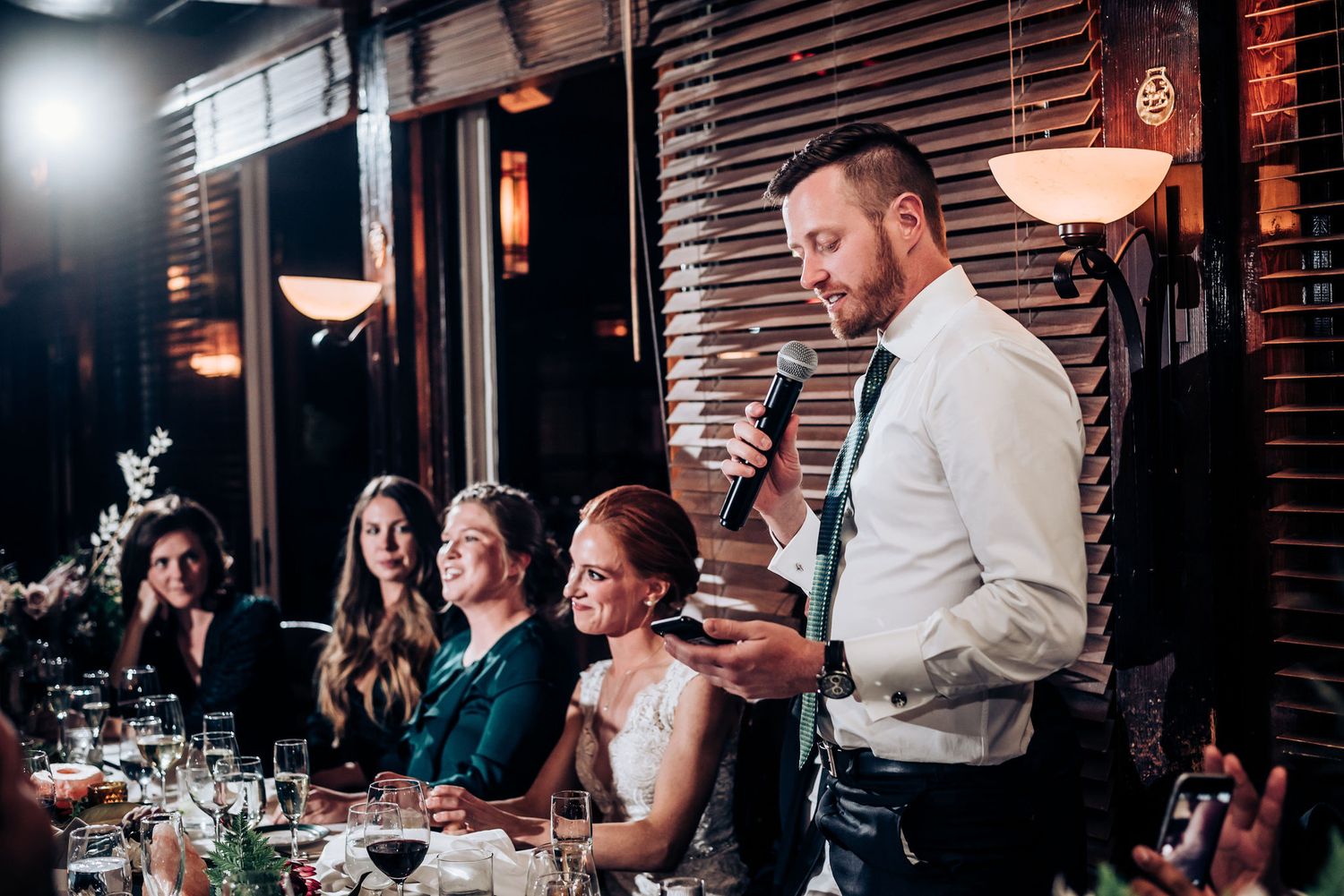 Wedding reception guests listen attentively to toasts and speeches in a dimly lit venue.