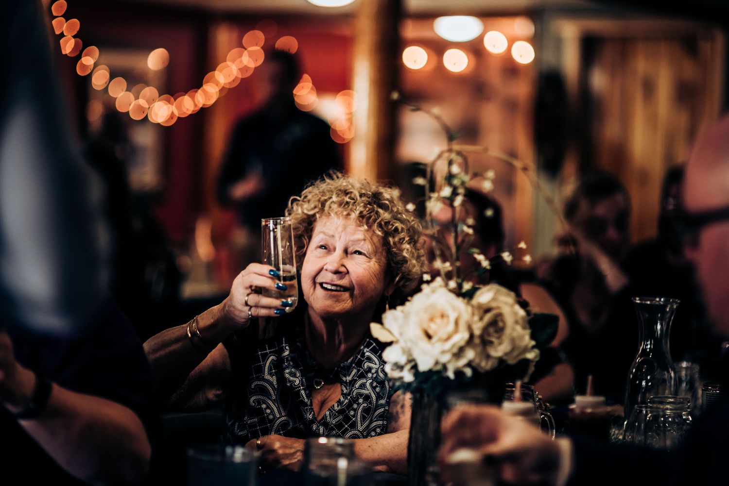 Wedding guest raising glass during reception with string lights and floral decorations in background.