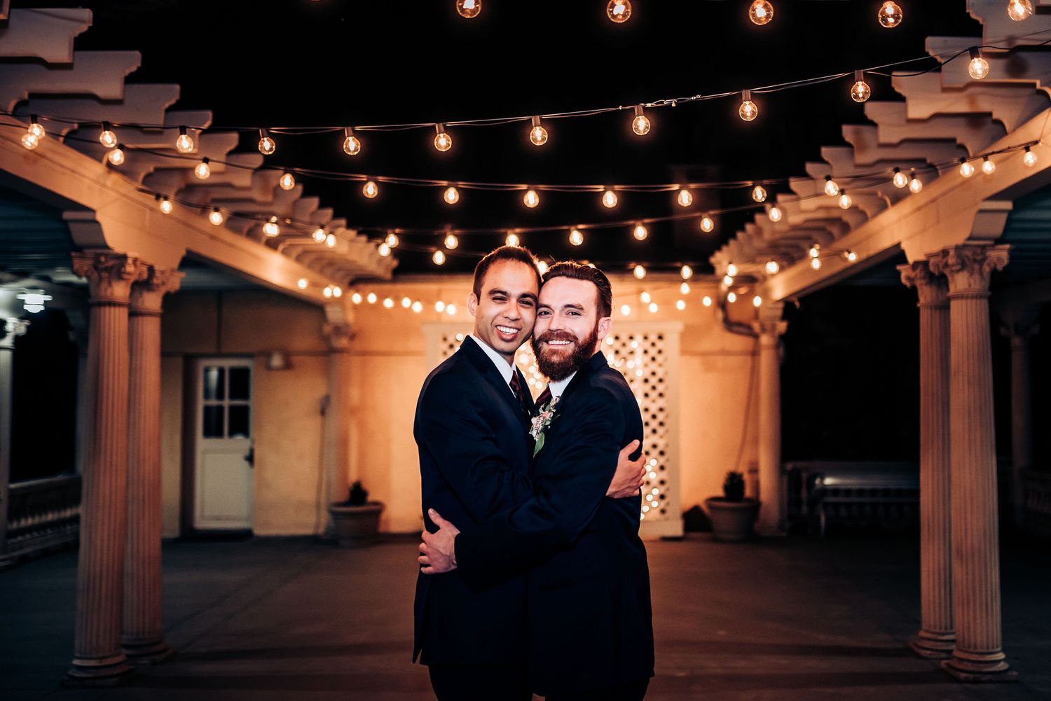Two people embrace under warm glowing string lights in an outdoor courtyard.