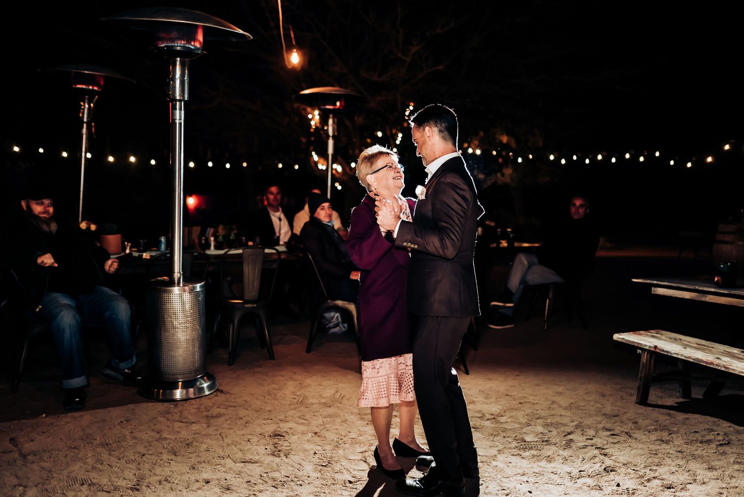 Couple shares a dance under outdoor string lights and patio heaters at evening event.