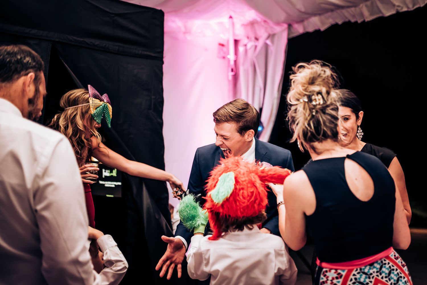 Wedding guests dancing and socializing at a reception with pink ambient lighting and black drapes.