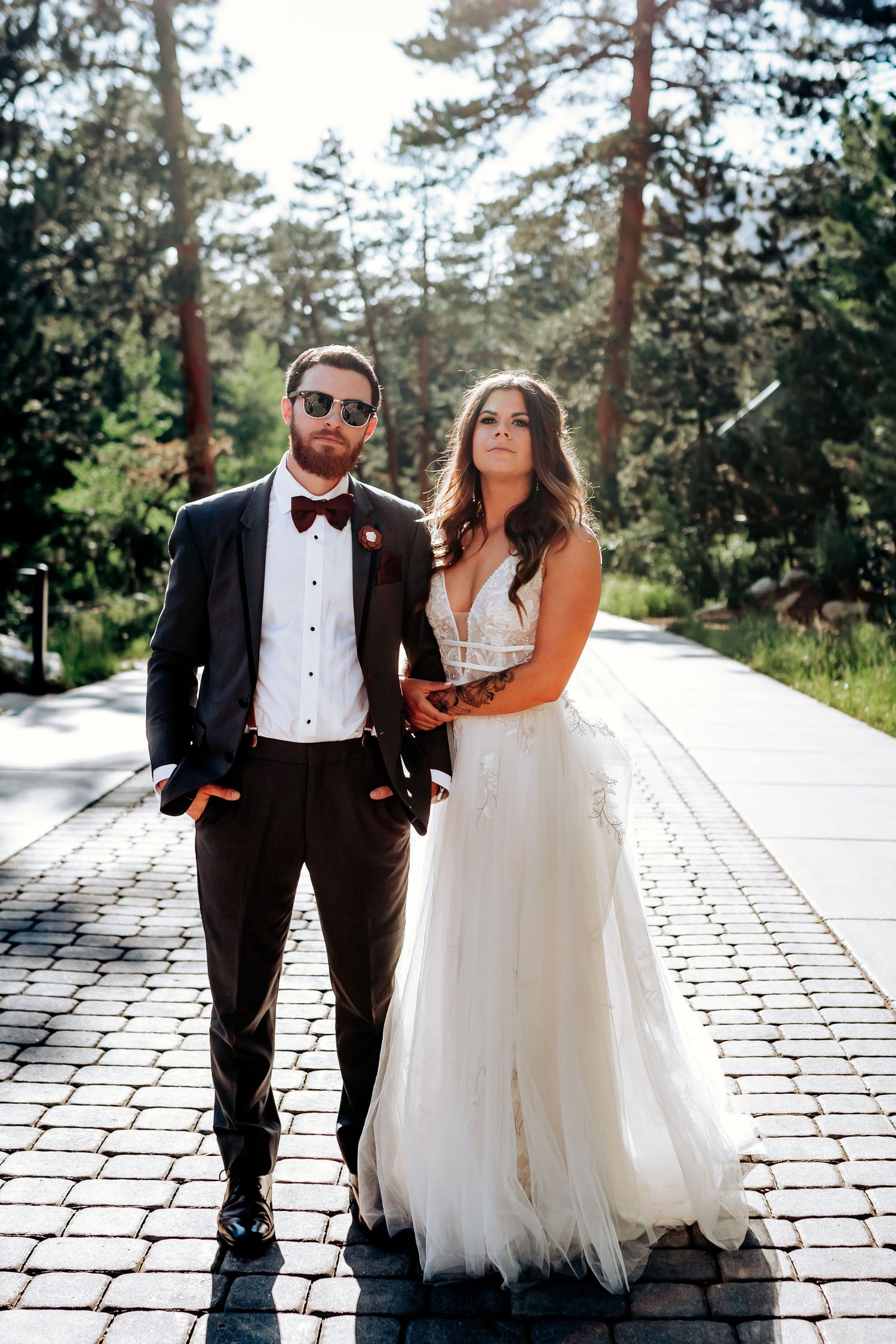A wedding couple poses on a stone path surrounded by tall pine trees in an outdoor forest setting.