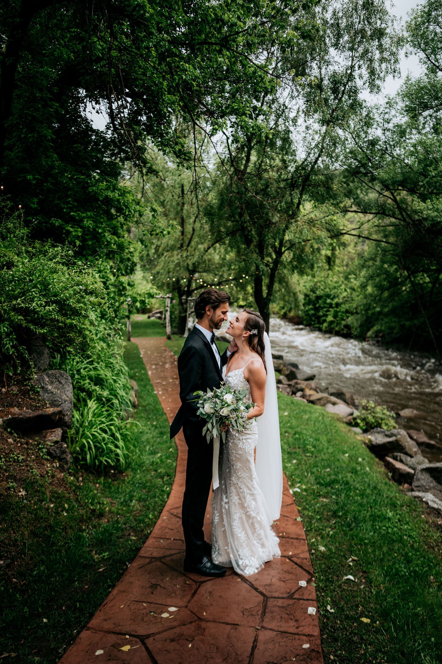 A romantic wedding couple embraces on a stone path beside a rushing stream in a lush green forest setting.