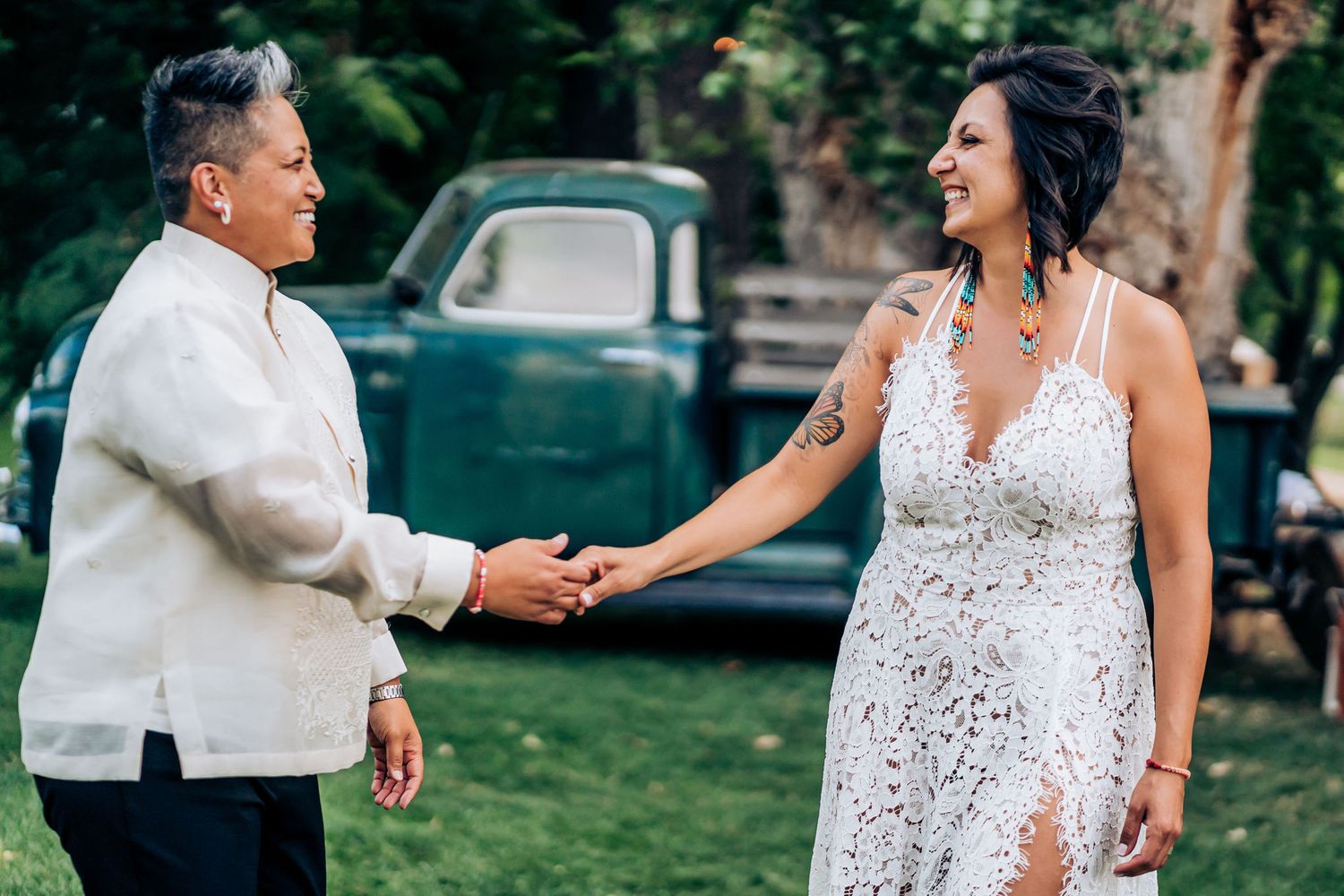 Couple in white formal wear holding hands outdoors with vintage truck in background.