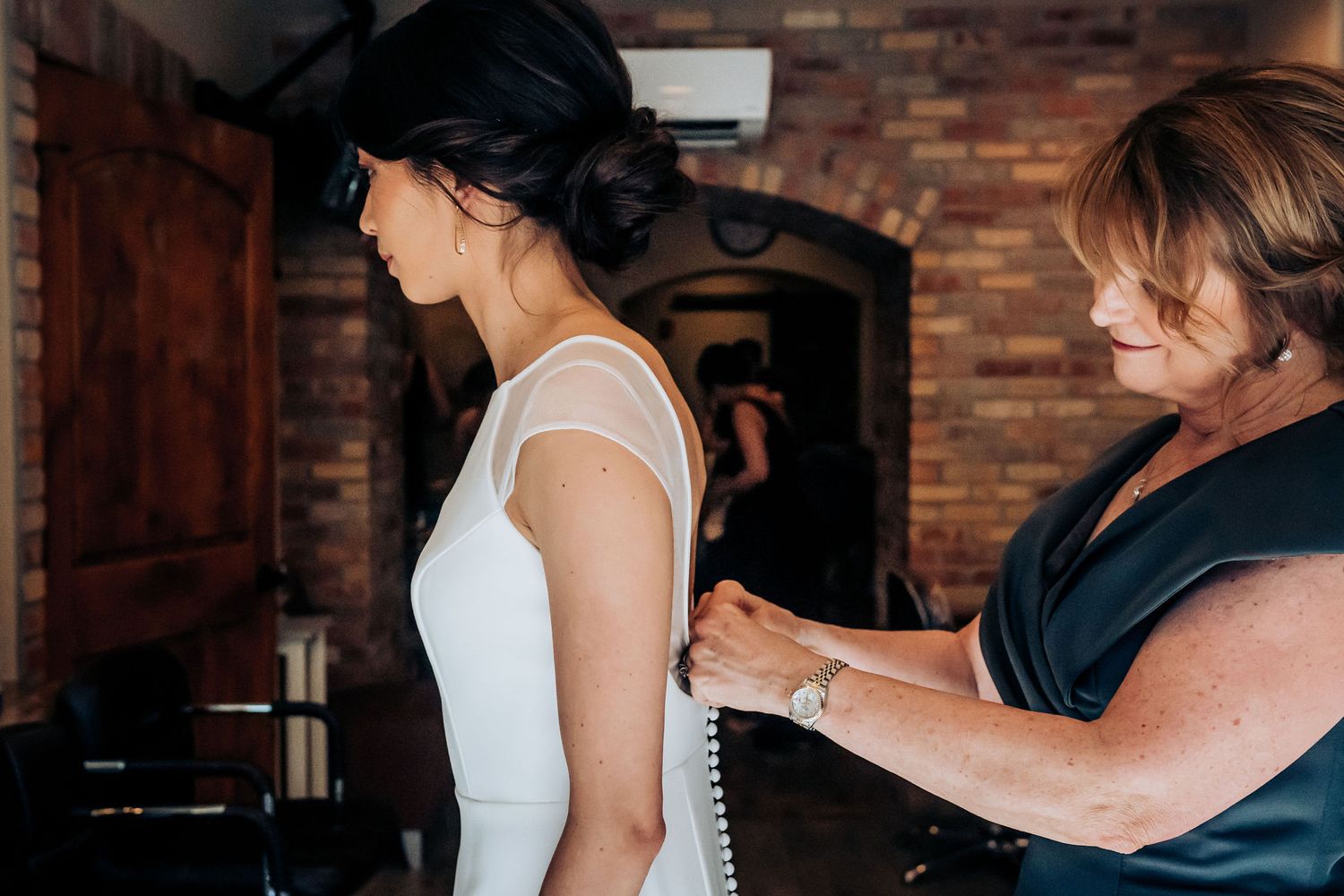 Final adjustments being made to a simple white wedding dress in a rustic venue setting.