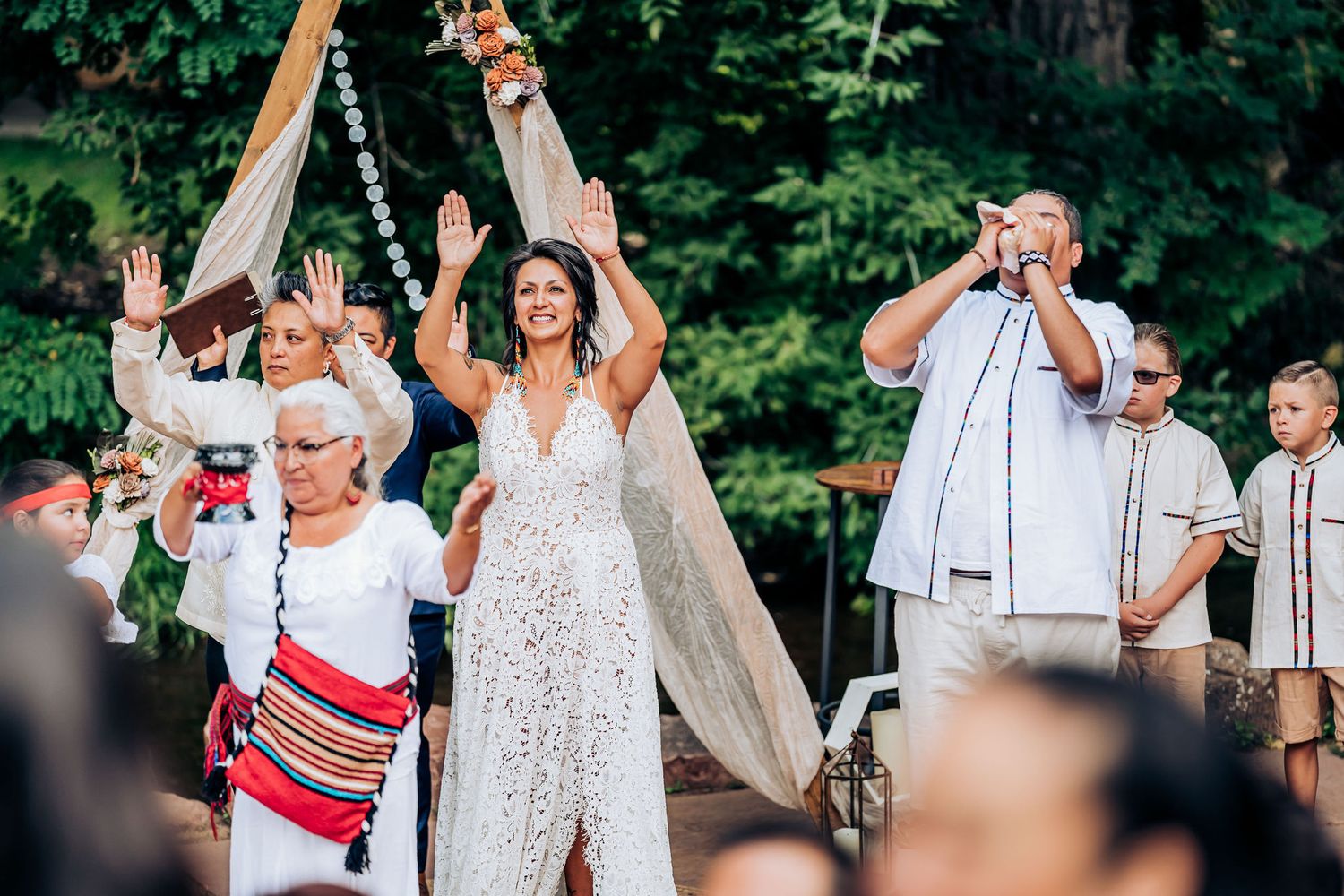 Celebratory outdoor wedding scene with people raising their hands under a wooden arch structure in a garden setting.