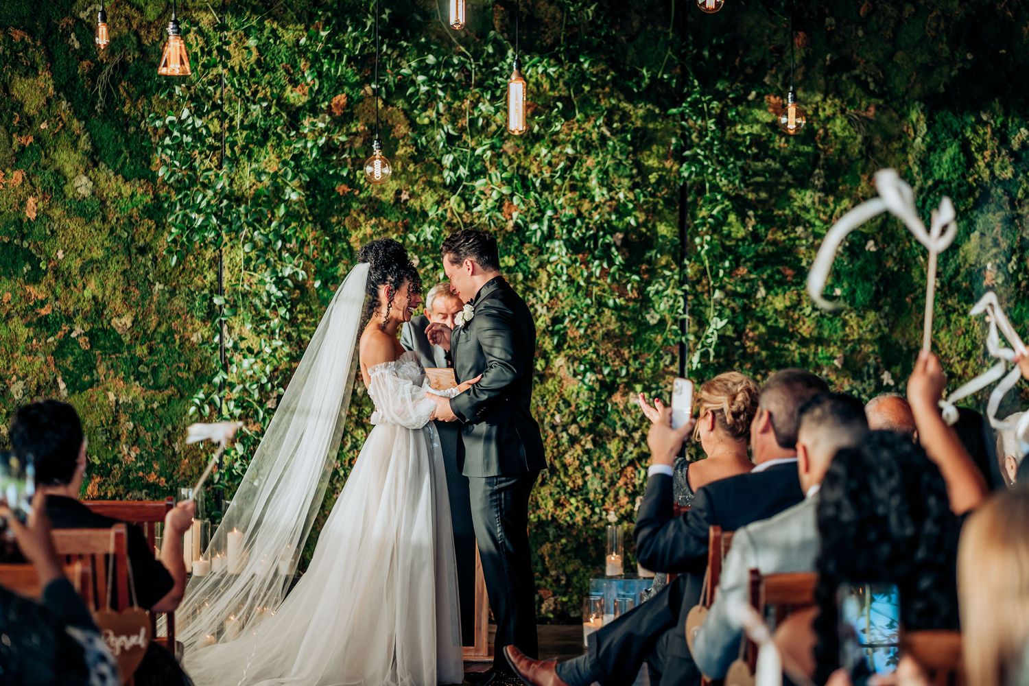Romantic ceremony moment against a garden wall with hanging lights and guests watching.