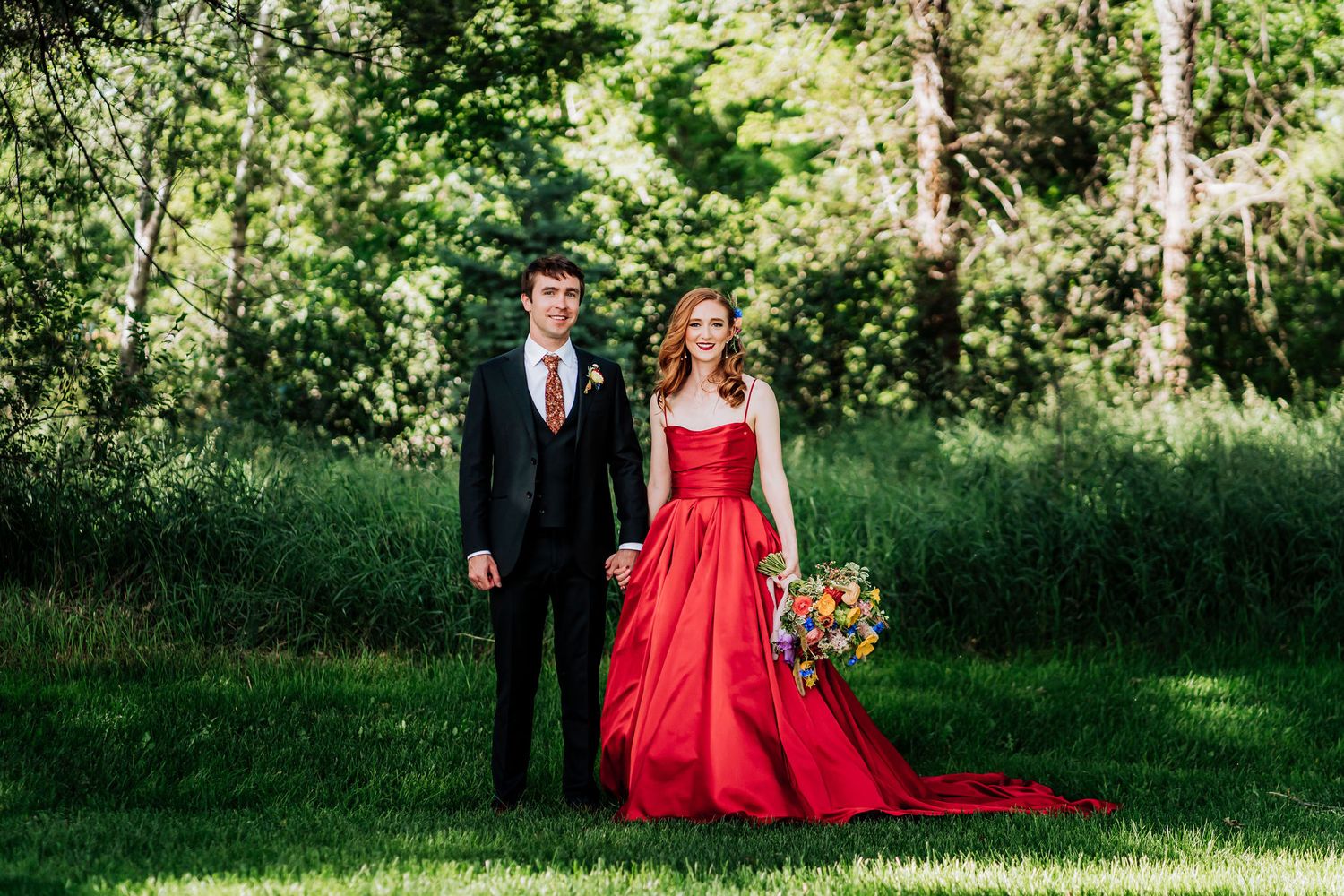 A formal couple poses in a garden setting, with her wearing a flowing red gown and him in a black suit.
