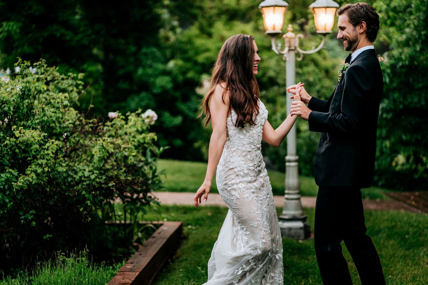 A couple poses near vintage lamp posts in a lush garden setting at dusk.