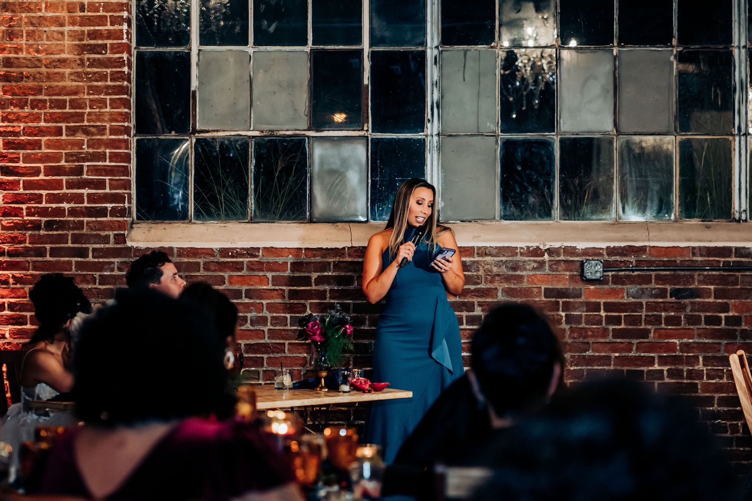 A speaker delivers a speech at a wedding reception against an exposed brick wall and industrial windows.