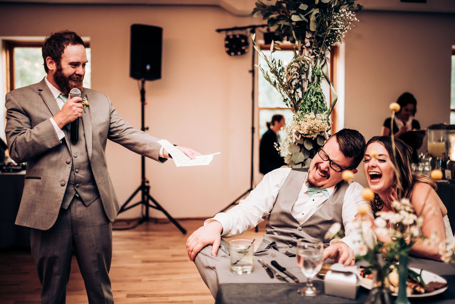 Wedding reception speech eliciting laughter from guests at indoor venue.