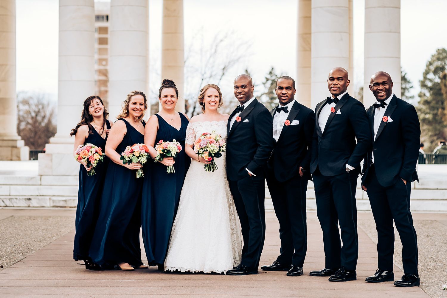 Wedding party in coordinating navy attire poses formally under classical architecture.