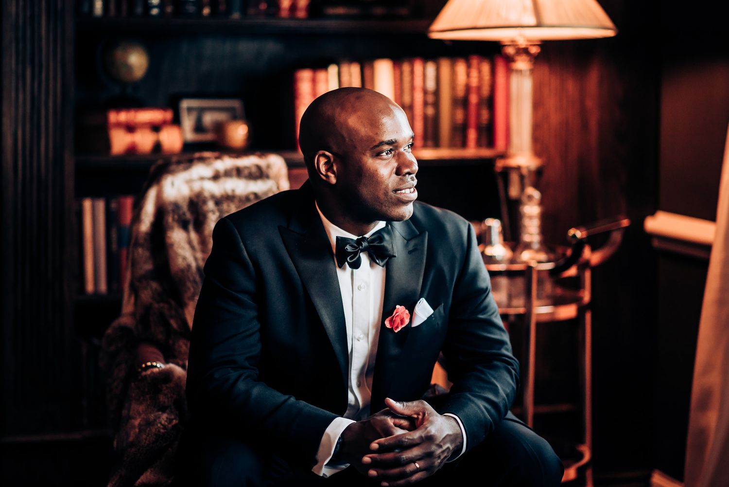 Contemplative groom in bow tie and tuxedo seated in elegant library setting.