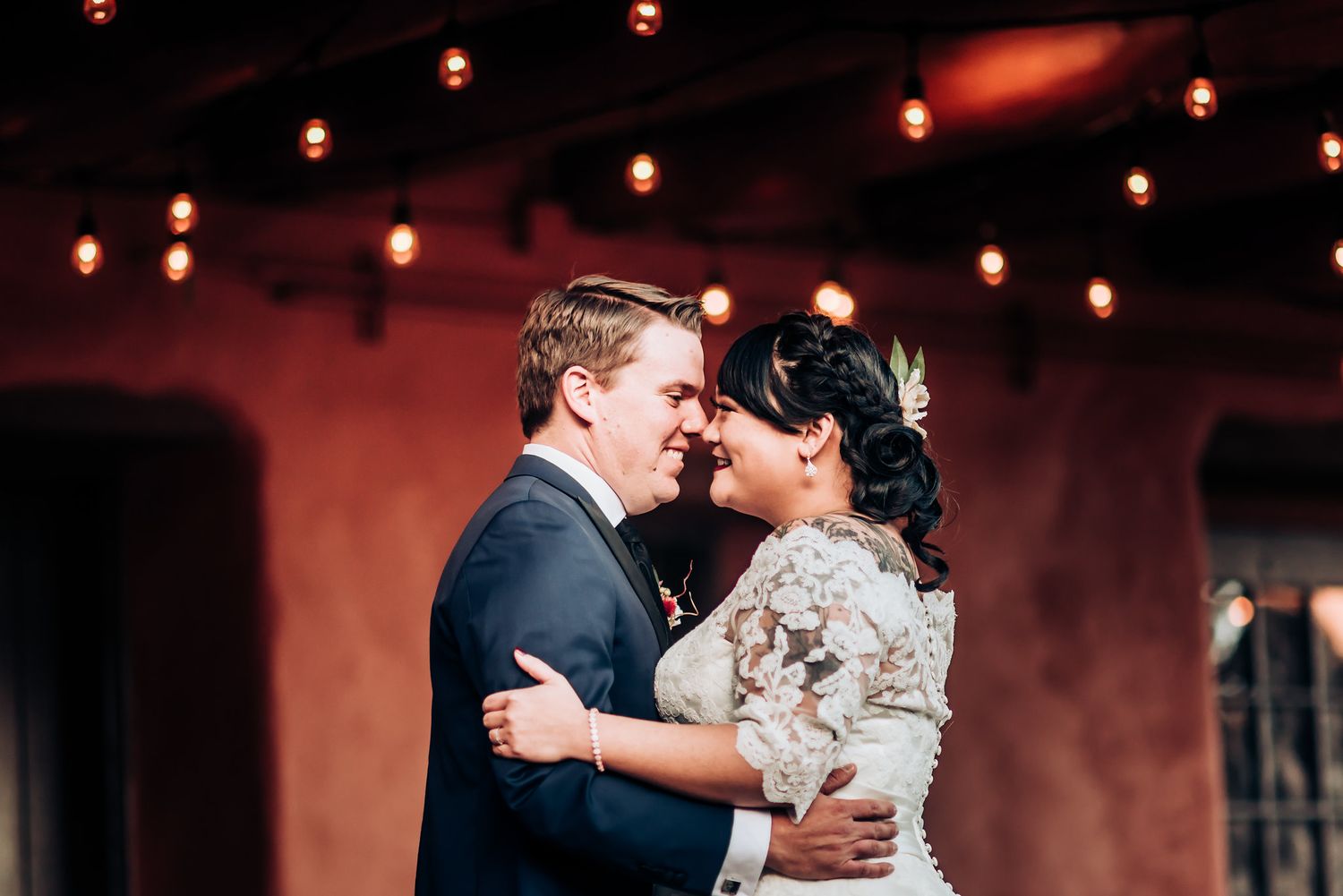 Wedding couple embracing under string lights against terra cotta backdrop.