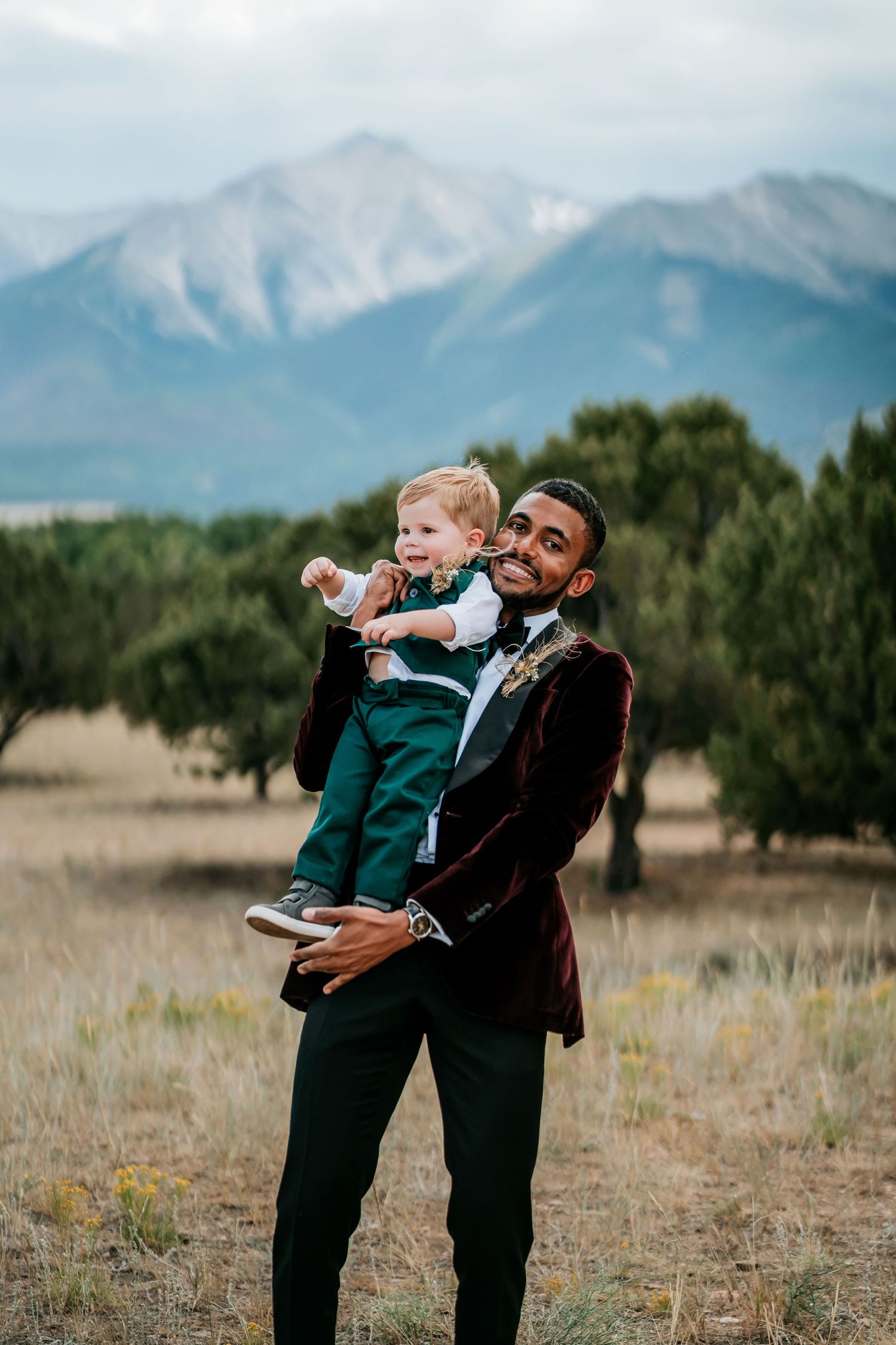 Person in burgundy velvet jacket holding small child against mountain backdrop.