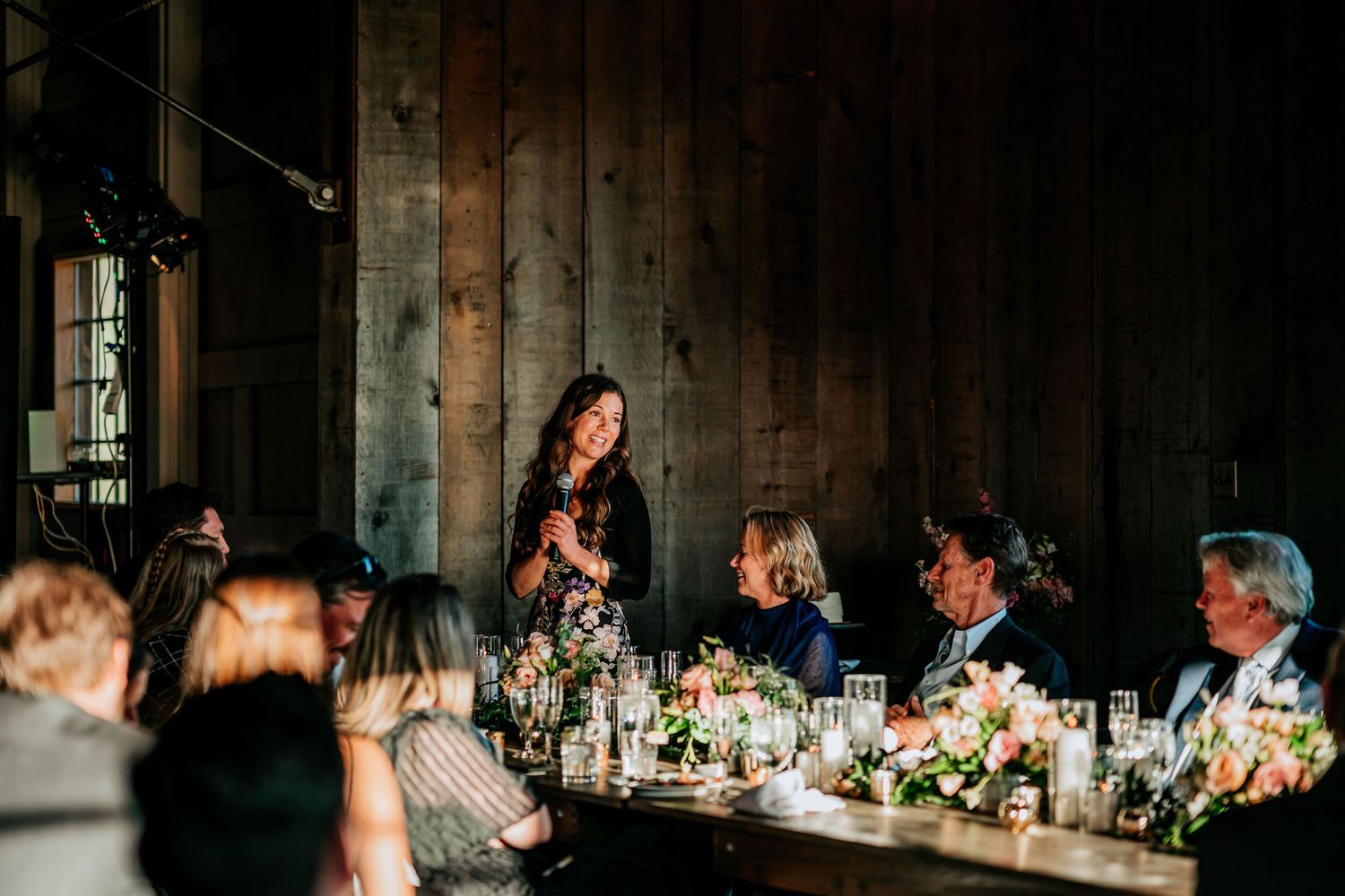 A wedding dinner reception in progress inside a dimly lit rustic barn with wooden walls and elegant table settings.