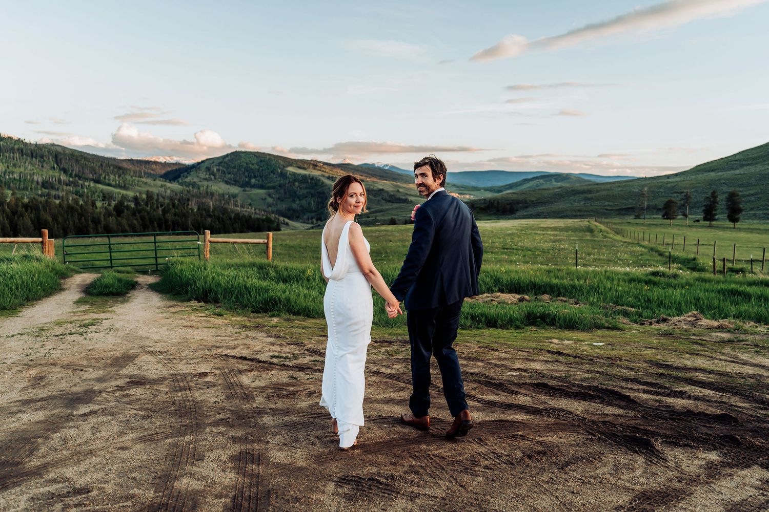 A couple in wedding attire stands on a dirt path with rolling green hills and mountains in the background.