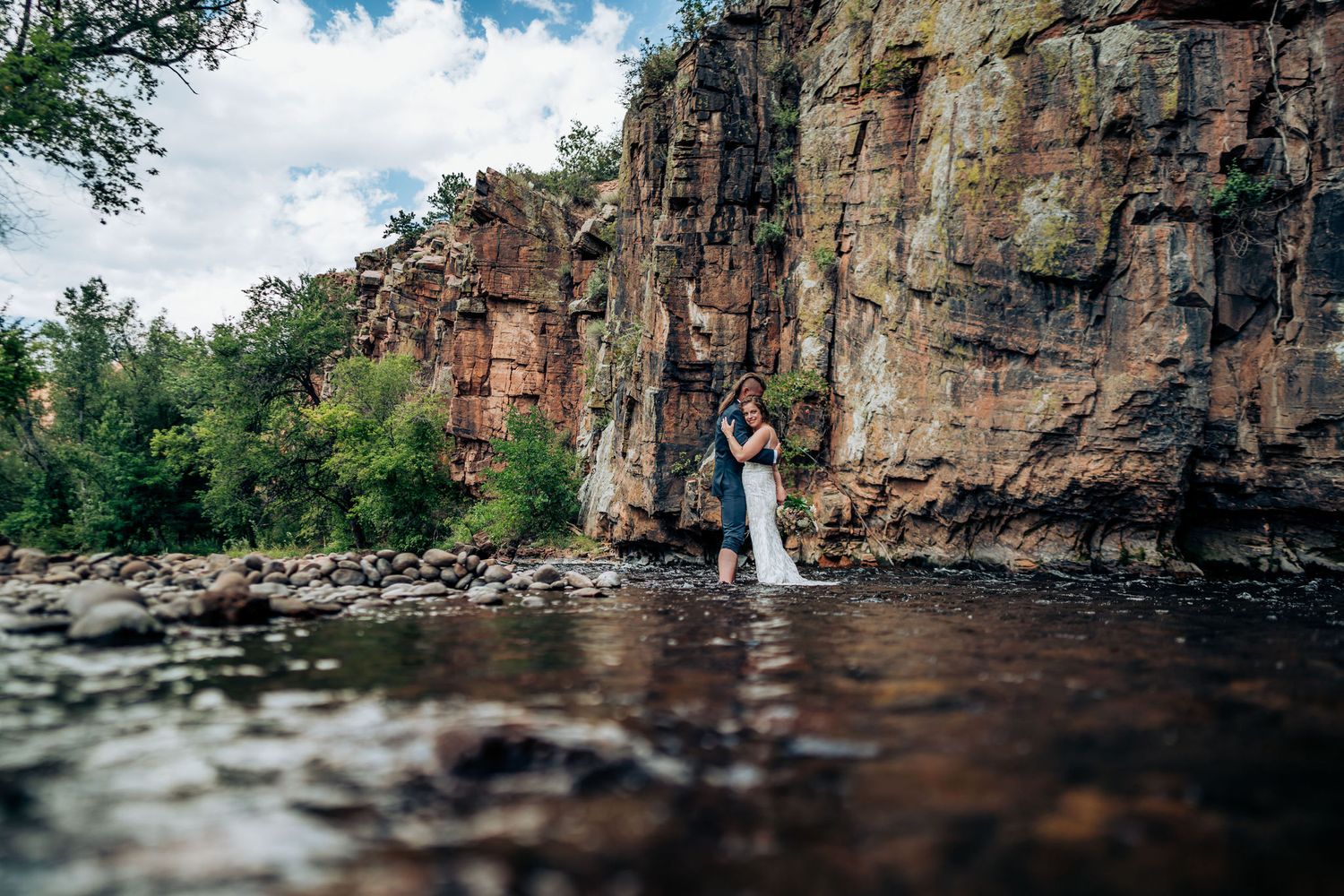 Wedding couple poses in shallow river water against dramatic red rock cliff wall.