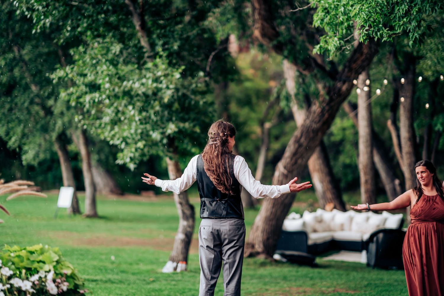 Person gesturing in a green outdoor wedding venue setting with tables visible in background.