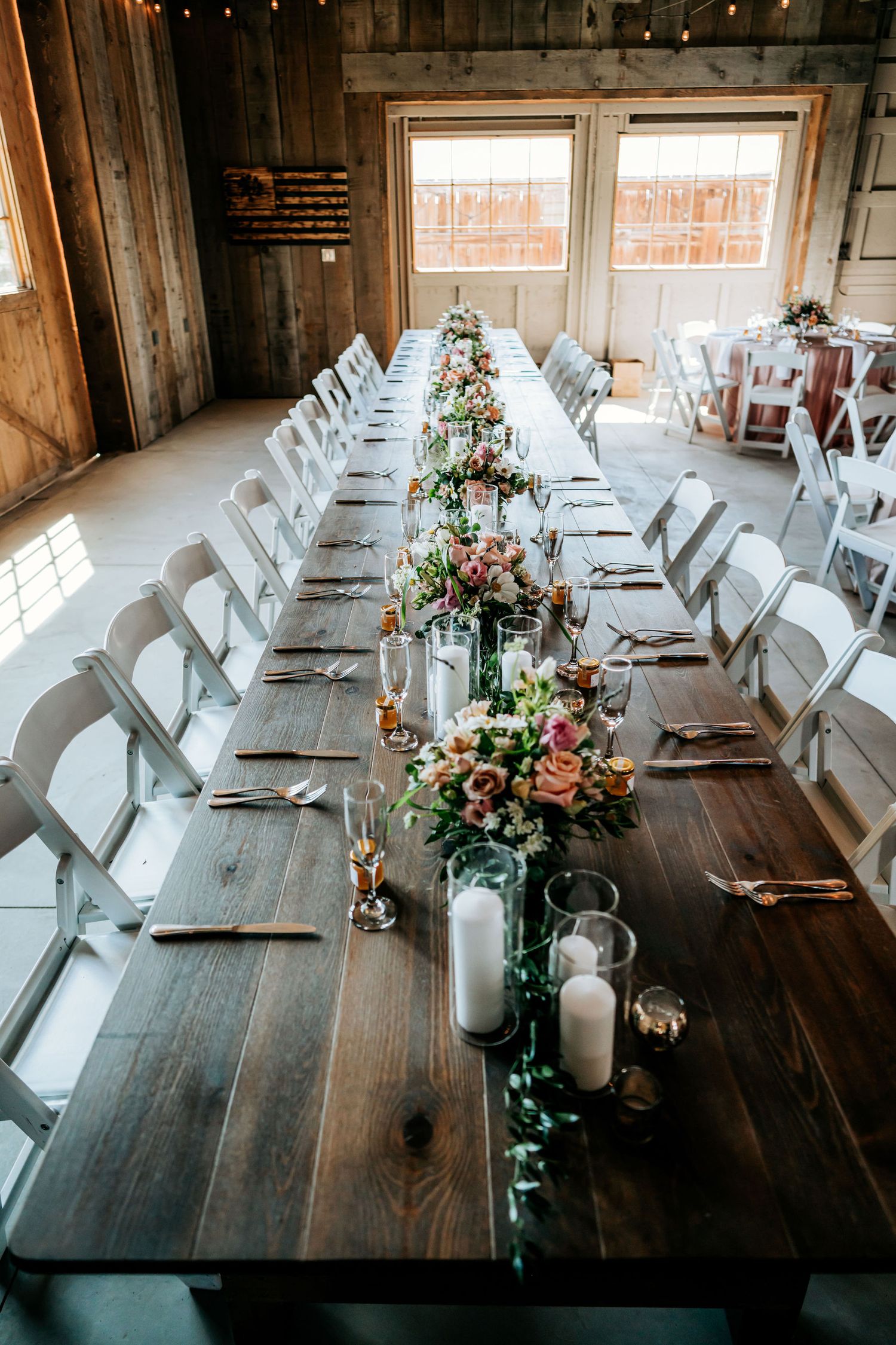 Long wooden farm table decorated with floral centerpieces and white chairs in barn venue.