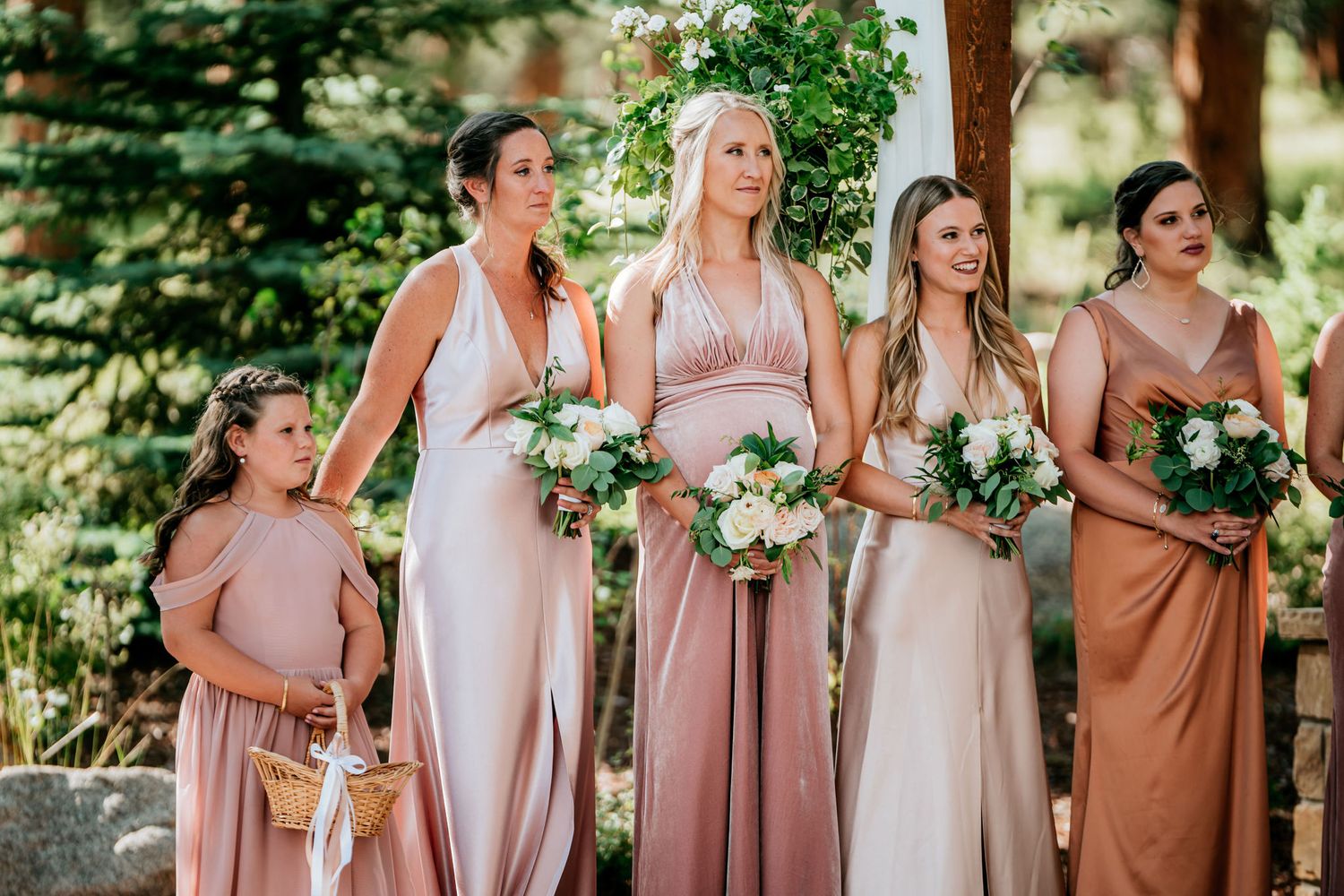 Bridesmaids in blush and neutral toned dresses stand together holding white bouquets.