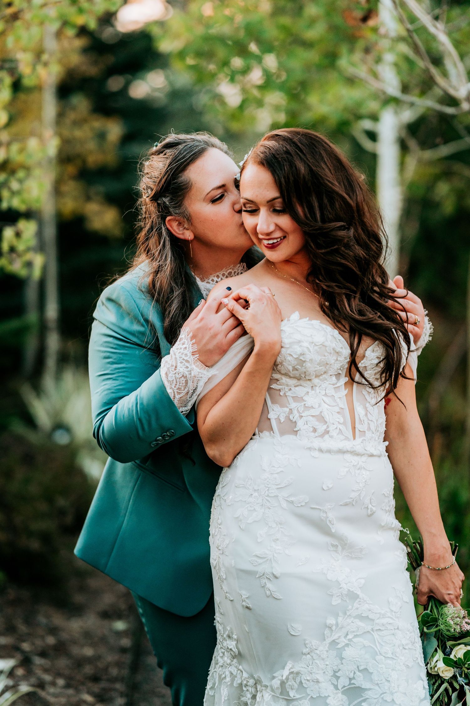 Bride in teal suit kissing partner in strapless lace mermaid wedding gown in garden setting.