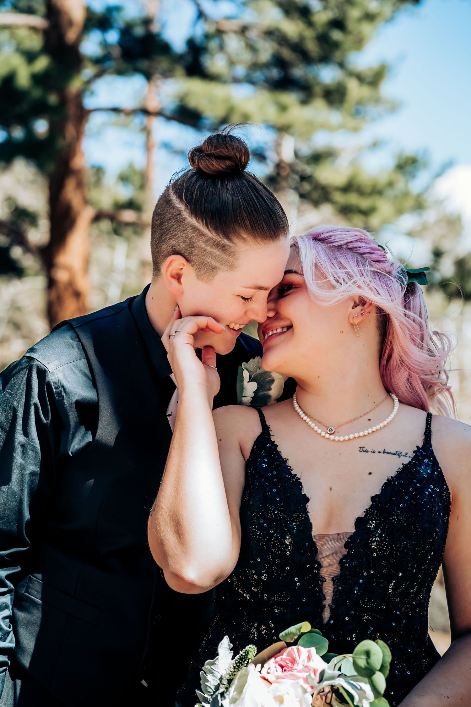Couple in formal attire shares playful moment against blue sky and pine trees.
